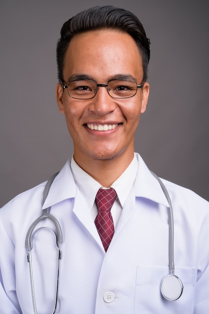 Young handsome Indian man doctor against gray wall 