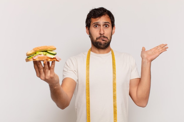 young handsome indian man doubting or uncertain expression and holding a sandwich