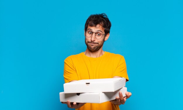 Young handsome indian man doubting or uncertain expression and holding take away pizzas