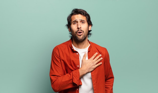 Young handsome indian man feeling shocked and surprised, smiling, taking hand to heart, happy to be the one or showing gratitude