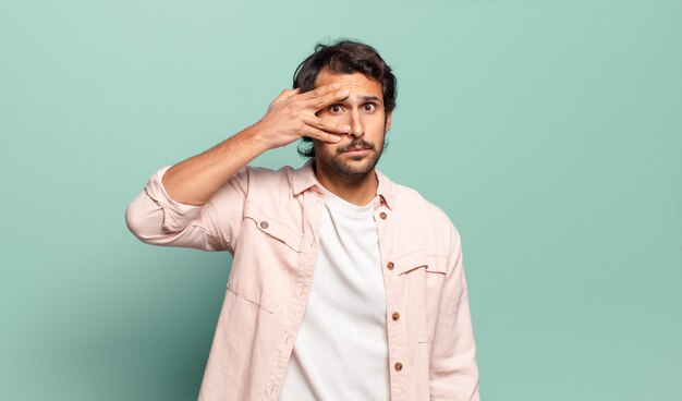 Young handsome indian man looking shocked, scared or terrified, covering face with hand and peeking between fingers