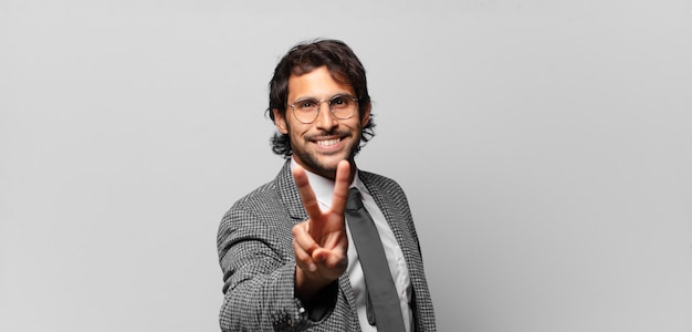 Young handsome indian man smiling and looking happy, carefree and positive, gesturing victory or peace with one hand. business concept