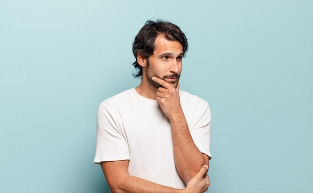 Young handsome indian man smiling with a happy, confident expression with hand on chin, wondering and looking to the side