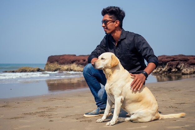 Young handsome indin man wearing black shirt and sunglasses,\
sitting on the beach with the white dog in goa beach happy morning\
time.pet travel puppy training