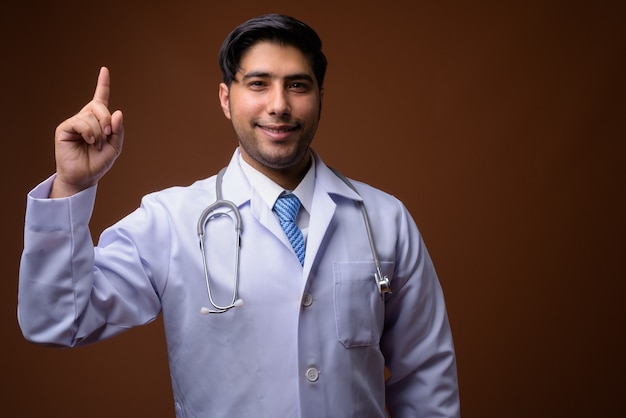 Young handsome Iranian man doctor against brown wall