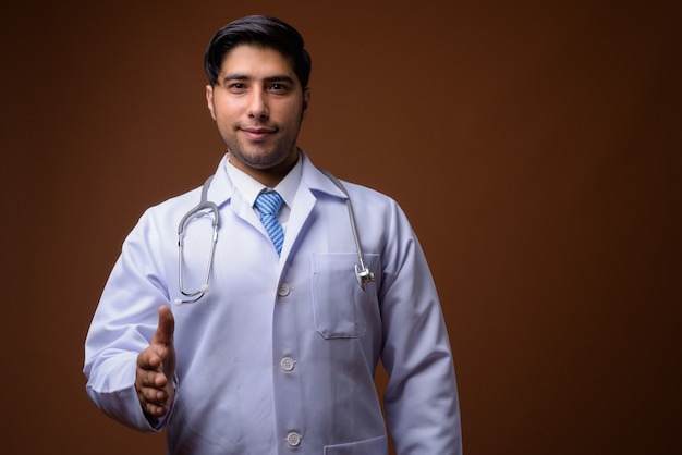 Young handsome Iranian man doctor against brown wall