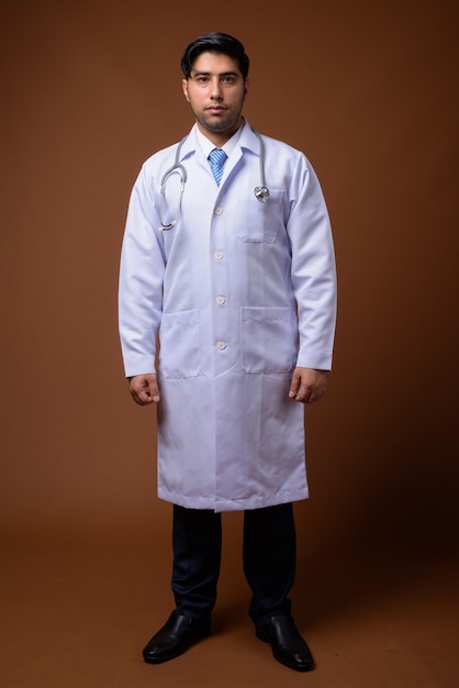 Young handsome Iranian man doctor against brown wall