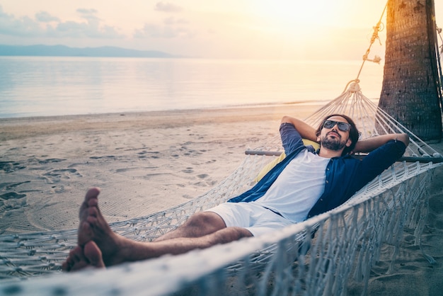Молодой красивый латинский человек в солнечных очках ослабляя в гамаке на пляже на заходе солнца на пляже.