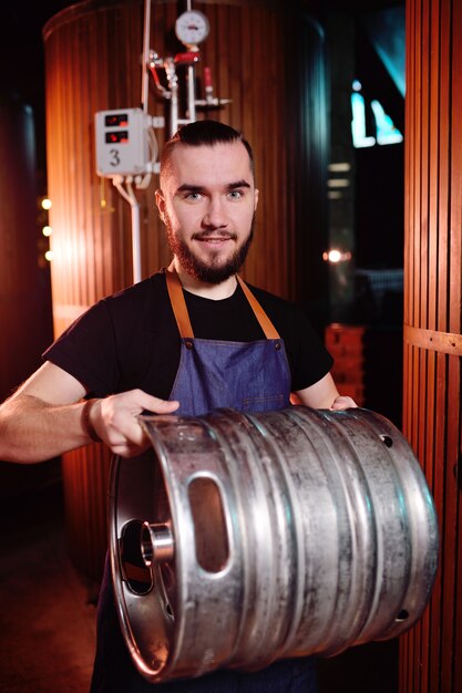 A young handsome male brewer holds an iron barrel with beer on the background of the brewery and beer tanks