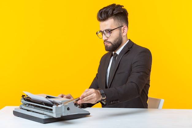 Молодой красивый мужчина-бизнесмен в формальной одежде, набирающий текст на пишущей машинке, позирует на желтом