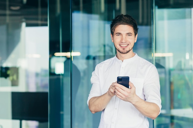 携帯電話を使用してオフィスに立っている若いハンサムな男性ビジネスマンはメッセージを収集します彼は立っているカメラの笑顔を見て