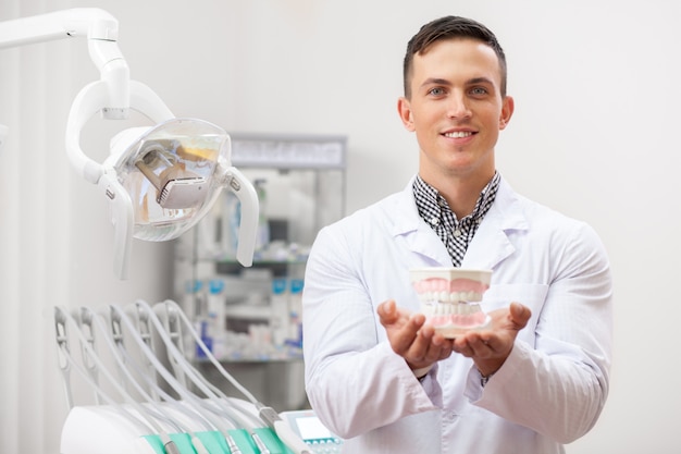 Young handsome male dentist smiling holding out denture mold to the camera