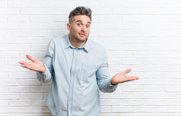 Young handsome man against a bricks wall doubting and shrugging shoulders in questioning gesture.