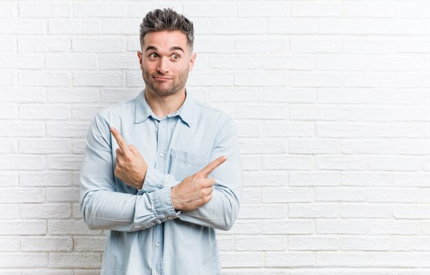 Young handsome man against a bricks wall points sideways, is trying to choose between two options.