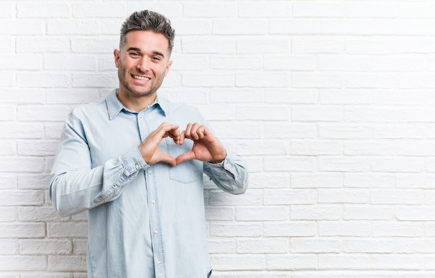 Young handsome man against a bricks wall smiling and showing a heart shape with hands.