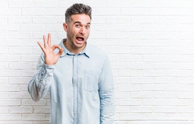 Il giovane uomo bello contro un muro di mattoni strizza l'occhio e tiene un gesto giusto con la mano.