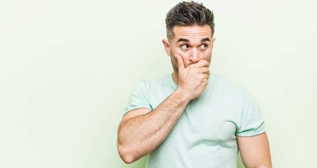 Young handsome man against a green wall thoughtful looking to a copy space covering mouth with hand.