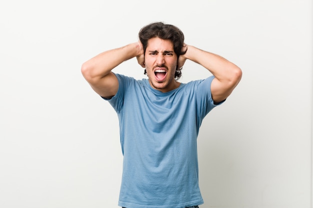 Young handsome man against a white wall covering ears with hands trying not to hear too loud sound.