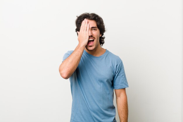 Young handsome man against a white wall having fun covering half of face with palm.