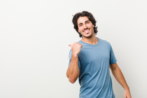 Young handsome man against a white wall points with thumb finger away, laughing and carefree.
