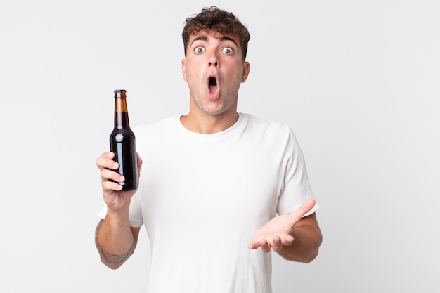 Young handsome man amazed, shocked and astonished with an unbelievable surprise and holding a beer bottle