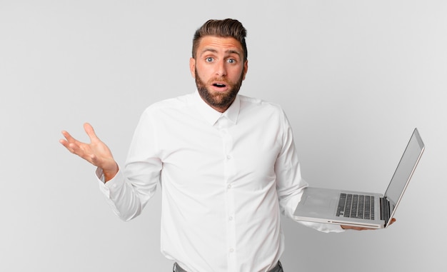 young handsome man amazed, shocked and astonished with an unbelievable surprise and holding a laptop