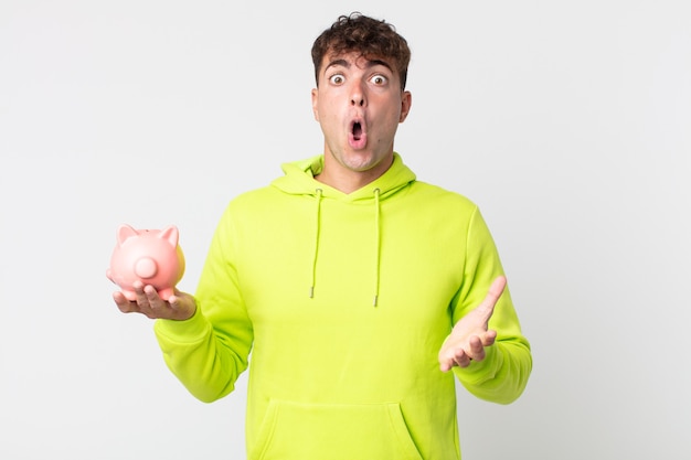Young handsome man amazed, shocked and astonished with an unbelievable surprise and holding a piggy bank