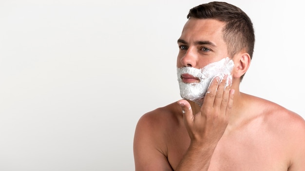 Young handsome man applying shaving cream standing against white wall