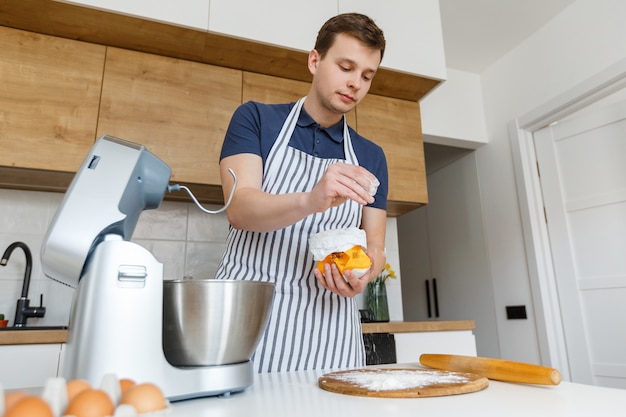 Giovane uomo bello in grembiule che setaccia la farina in cucina concetto di cibo fatto in casa stile di vita domestico