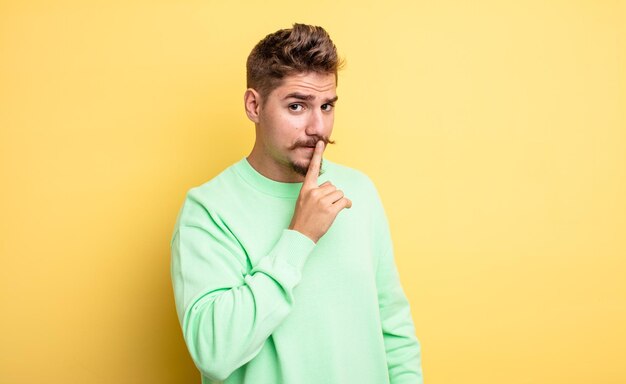 Young handsome man asking for silence and quiet, gesturing with finger in front of mouth, saying shh or keeping a secret. strange moustache concept