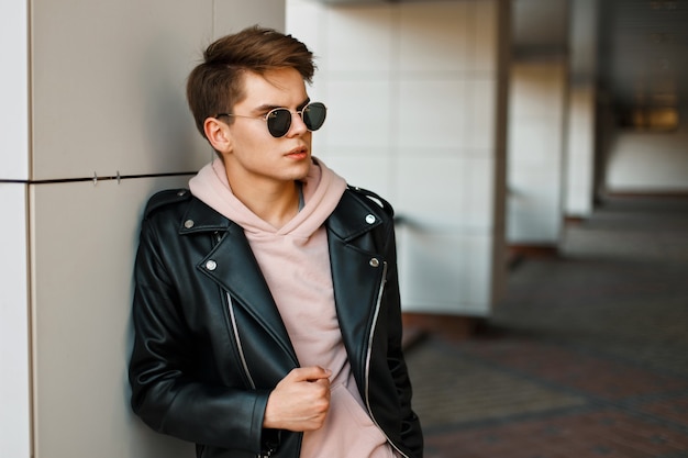 Young handsome man in aviator sunglasses in a black leather jacket and a pink sweatshirt near a modern white wall