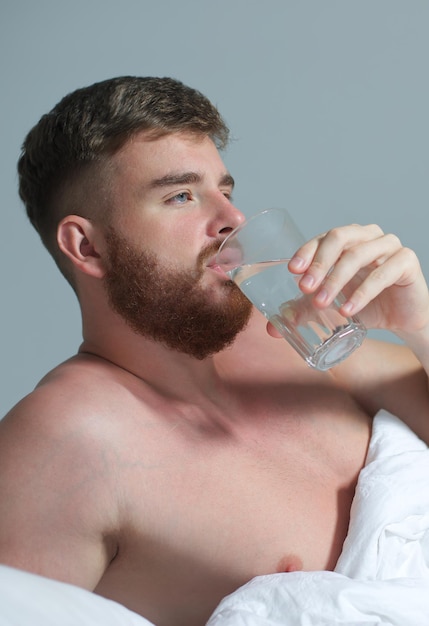 朝、自宅の寝室のベッドで若いハンサムな男が白い毛布の下に横たわり、ガラスから純粋な新鮮な水を飲む
