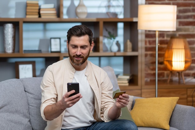 A young handsome man in a beige shirt is sitting on the sofa at home using a phone and a credit card