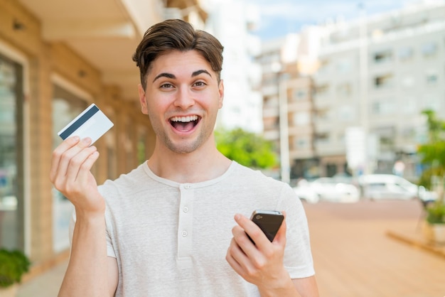 Молодой красивый мужчина покупает с помощью мобильного телефона и держит кредитную карту с удивленным выражением лица