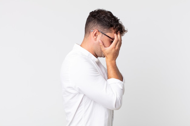 Young handsome man covering eyes with hands with a sad, frustrated look of despair, crying, side view