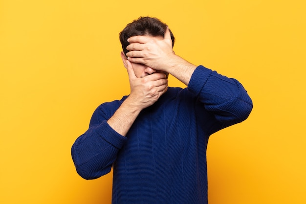 Young handsome man covering face with both hands saying no to the camera! refusing pictures or forbidding photos