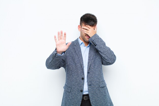 Young handsome man covering face with hand and putting other hand up front to stop, refusing photos or pictures against white wall