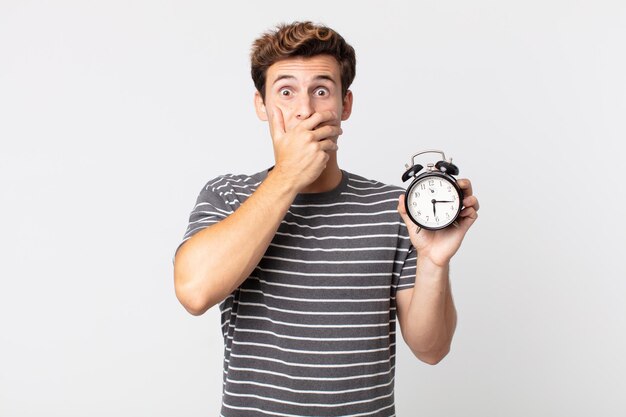 Young handsome man covering mouth with hands with a shocked and holding an alarm clock