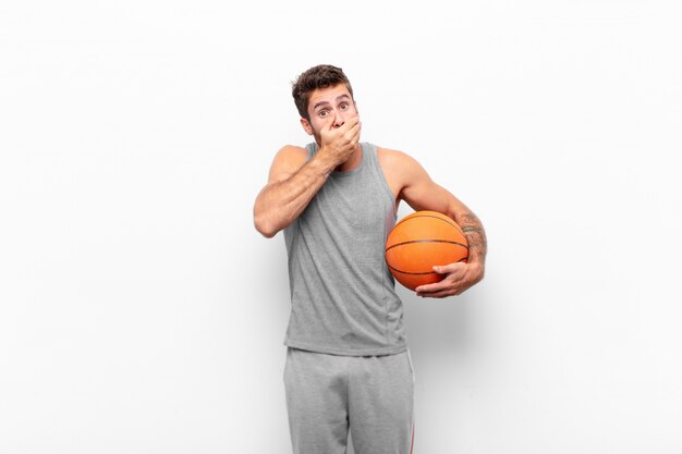 Young handsome man covering mouth with hands with a shocked, surprised expression, keeping a secret or saying oops holding a basketball ball.