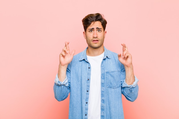 Young handsome man crossing fingers anxiously and hoping for good luck with a worried look against pink wall