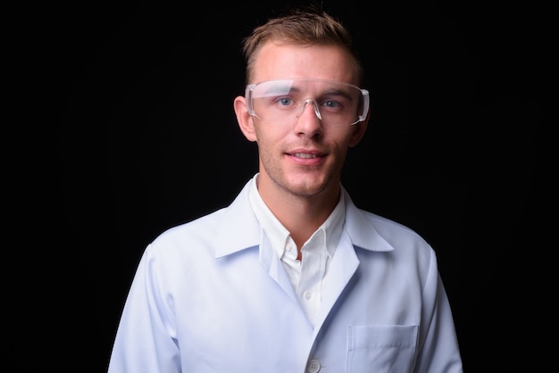  young handsome man doctor with blond hair wearing protective glasses against black wall