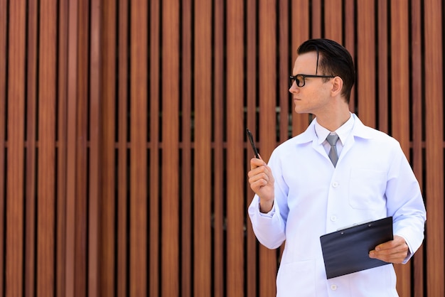 Young handsome man doctor with eyeglasses holding clipboard outdoors