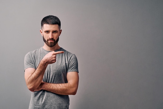 A young handsome man dressed in a gray Tshirt and light jeans looks into the camera showing direction and pointing with finger Copy space