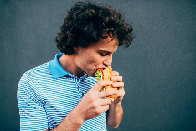 健康的なハンバーガーを食べる若いハンサムな男屋外でハンバーガーを食べるファーストフードレストランで空腹の男ストリートフードを食べてハンバーガーを食べる巻き毛の男