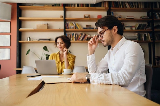 Young handsome man in eyeglasses sitting at the table thoughtfully working with colleague on background in modern office