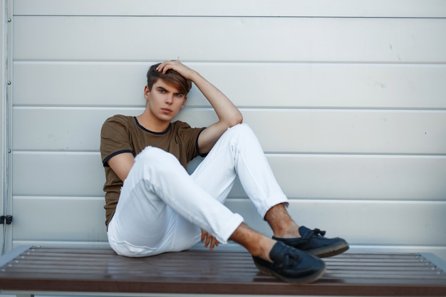Young handsome man in fashionable clothes sitting on a bench near a gray glossy wall
