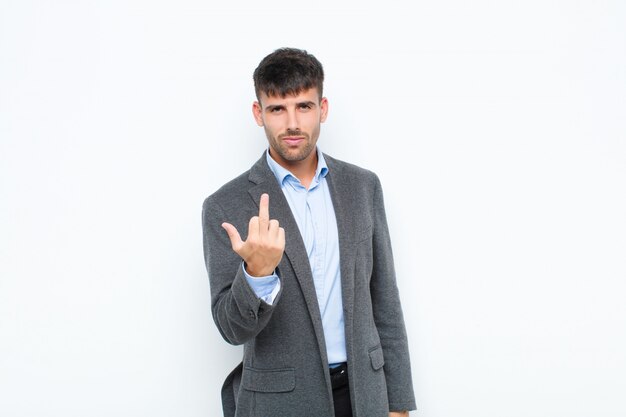 Young handsome man feeling angry, annoyed, rebellious and aggressive, flipping the middle finger, fighting back on white wall