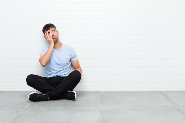 Young handsome man feeling bored, frustrated and sleepy after a tiresome, dull and tedious task, holding face with hand sitting on cement floor