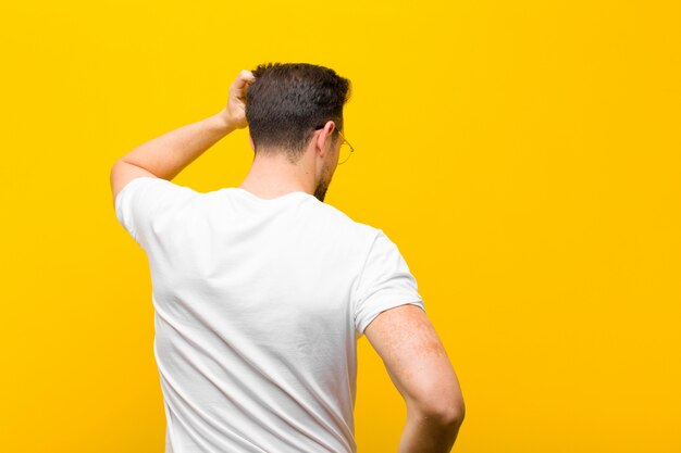 Photo young handsome man feeling clueless and confused, thinking a solution, with hand on hip and other on head, rear view against orange wall