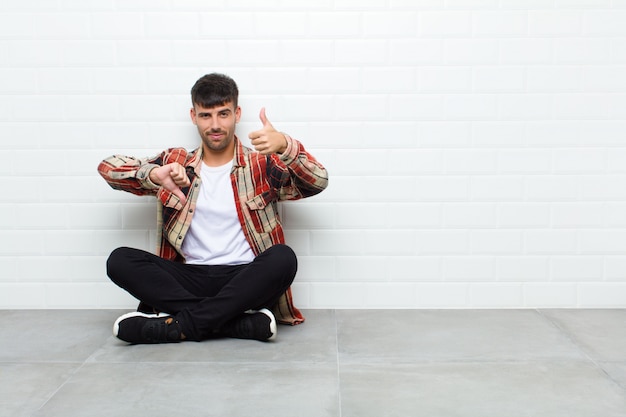 Young handsome man feeling confused, clueless and unsure,\
weighting the good and bad in different options or choices sitting\
on cement floor